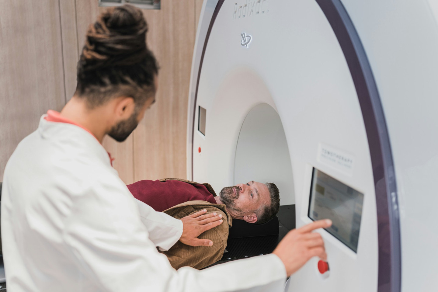 A patient lying on the couch of a radiotherapy machine, about to be moved into its ring-shaped gantry, while a radiographer steadies them with one hand and sets up the treatment on the control screen with the other